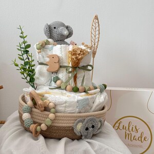 Op de afbeelding: Een baby shower cadeau mand met een neutrale kleurstelling. De mand is gevuld met luiers, een gehaakt olifantenspeelgoed, een houten bijtring en een houten rammelaar. De mand is versierd met gedroogde bloemen en een groene plant.