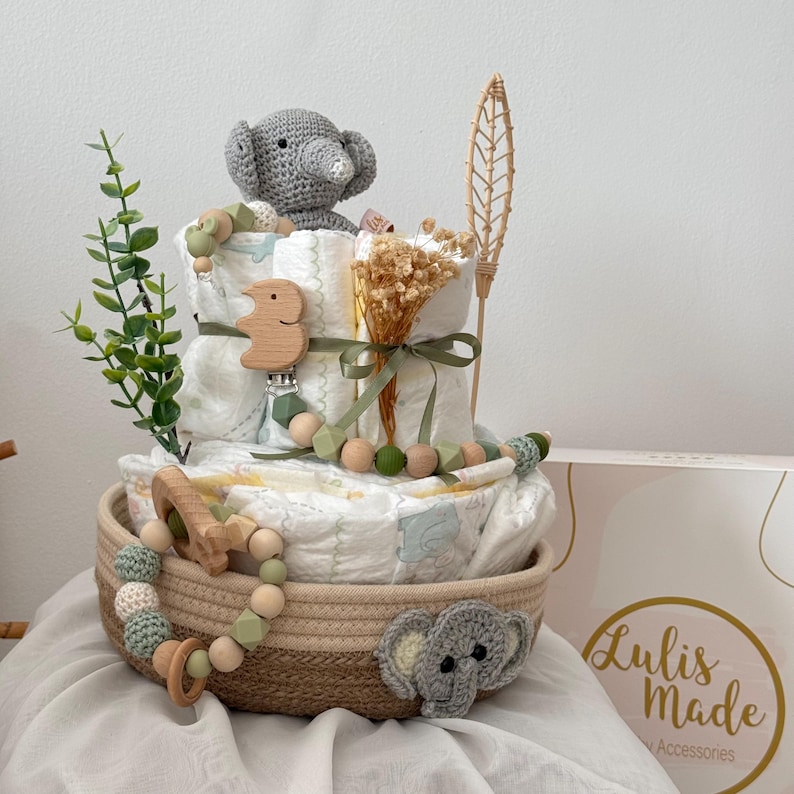 Op de afbeelding: Een baby shower cadeau mand met een neutrale kleurstelling. De mand is gevuld met luiers, een gehaakt olifantenspeelgoed, een houten bijtring en een houten rammelaar. De mand is versierd met gedroogde bloemen en een groene plant.