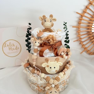 K&ouml;nnte beinhalten: Ein dreistufiger Babygeschenkkorb mit Giraffen-Motiv. Der Korb ist mit geh&auml;kelten Giraffenk&ouml;pfen, Holzperlen und einem braunen Band verziert. Der Korb enth&auml;lt Windeln, einen Schnuller und Gr&uuml;nzeug. Die Box im Hintergrund tr&auml;gt den Text "Lulis Made".