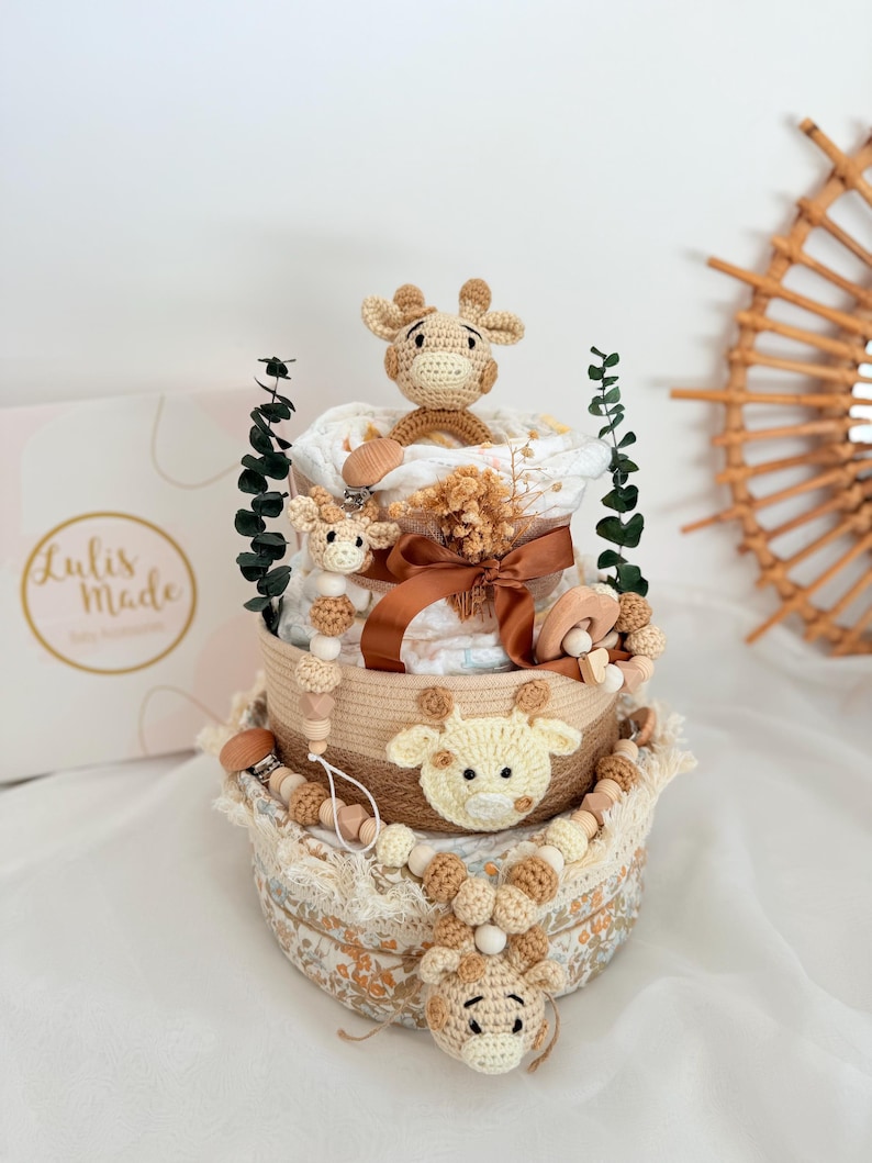 K&ouml;nnte beinhalten: Ein dreistufiger Babygeschenkkorb mit Giraffen-Motiv. Der Korb ist mit geh&auml;kelten Giraffenk&ouml;pfen, Holzperlen und einem braunen Band verziert. Der Korb enth&auml;lt Windeln, einen Schnuller und Gr&uuml;nzeug. Die Box im Hintergrund tr&auml;gt den Text "Lulis Made".