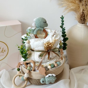 May include: A diaper cake with two tiers, decorated with a green crocheted turtle, wooden toys, and greenery. The cake sits in a woven basket and is adorned with a ribbon. A box with the word "ide" is visible in the background.
