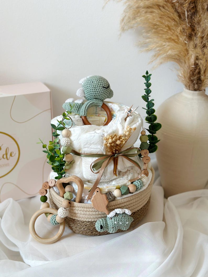 May include: A diaper cake with two tiers, decorated with a green crocheted turtle, wooden toys, and greenery. The cake sits in a woven basket and is adorned with a ribbon. A box with the word "ide" is visible in the background.