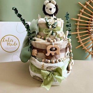 May include: A three-tiered diaper cake decorated with green ribbons, wooden beads, and a crocheted lion topper. The cake features wooden elephant and hedgehog accents, and is adorned with eucalyptus branches. The box in the background has the text "Lulis Made Baby Accessories".