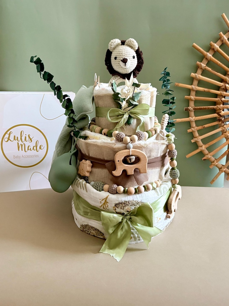 May include: A three-tiered diaper cake decorated with green ribbons, wooden beads, and a crocheted lion topper. The cake features wooden elephant and hedgehog accents, and is adorned with eucalyptus branches. The box in the background has the text "Lulis Made Baby Accessories".