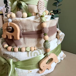 May include: A multi-tiered baby gift basket with a light green ribbon and wooden elephant-shaped teething toys. The basket is decorated with green and brown accents, and includes a variety of baby items, such as blankets and toys.