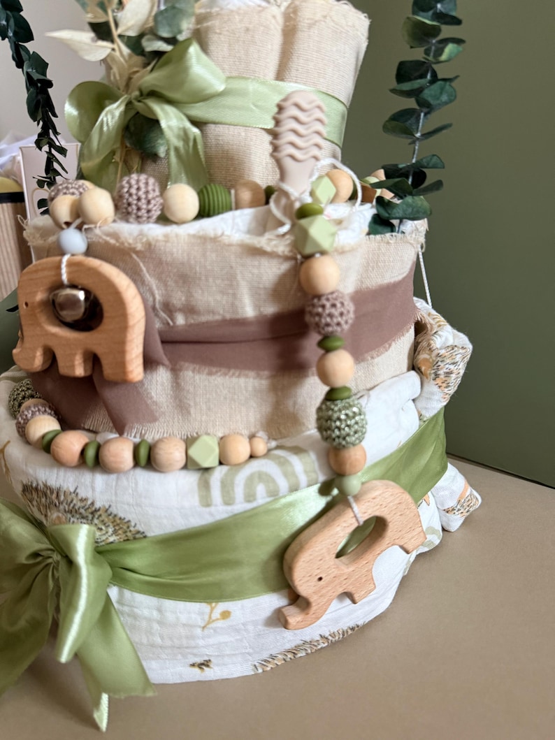 May include: A multi-tiered baby gift basket with a light green ribbon and wooden elephant-shaped teething toys. The basket is decorated with green and brown accents, and includes a variety of baby items, such as blankets and toys.
