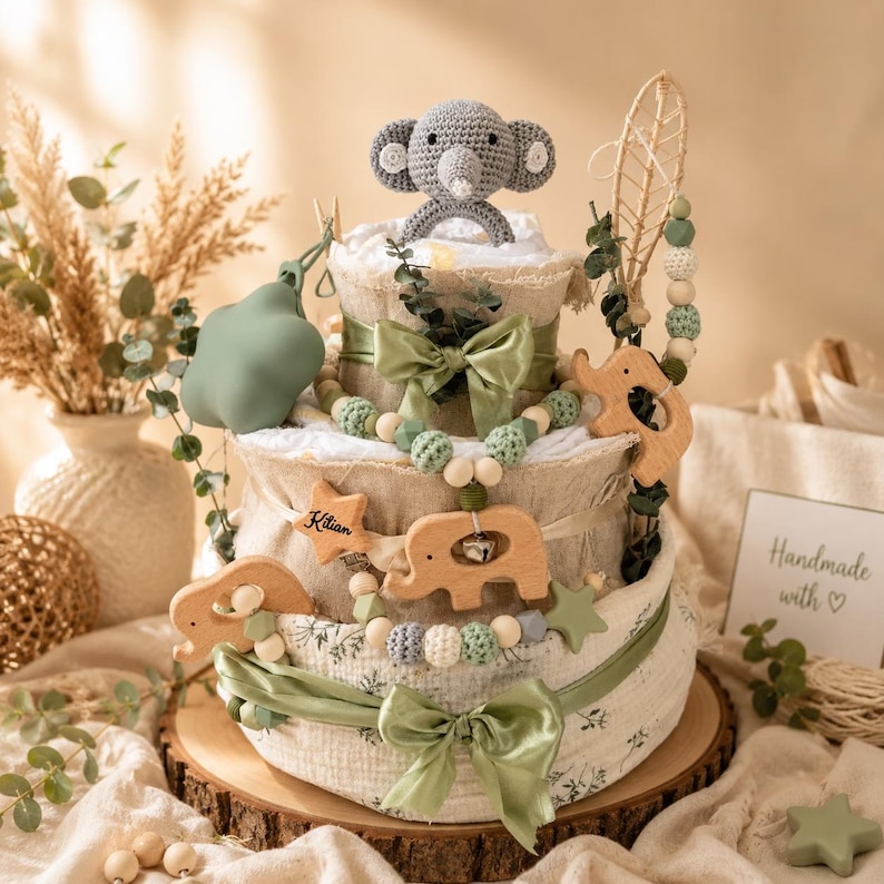 May include: A three-tiered baby gift cake with a gray crocheted elephant topper. The cake is decorated with green bows, wooden elephant and star accents, and a beaded garland. The base is on a wooden round. The words "Handmade with ♡" are on a card.