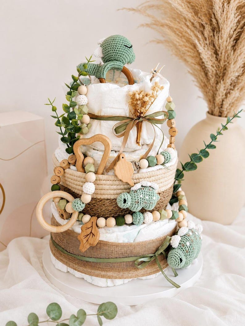 May include: A diaper cake with three tiers, decorated with green and beige accents. The cake features a crocheted dinosaur, wooden beads, and a wooden ring. The cake is adorned with greenery and a ribbon, creating a charming baby shower gift.