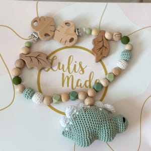 May include: A baby pacifier clip with a green crocheted dinosaur, wooden beads, and leaf accents. The clip features a wooden butterfly-shaped clip and the text "Lulis Made". The color palette includes shades of green, white, and natural wood.