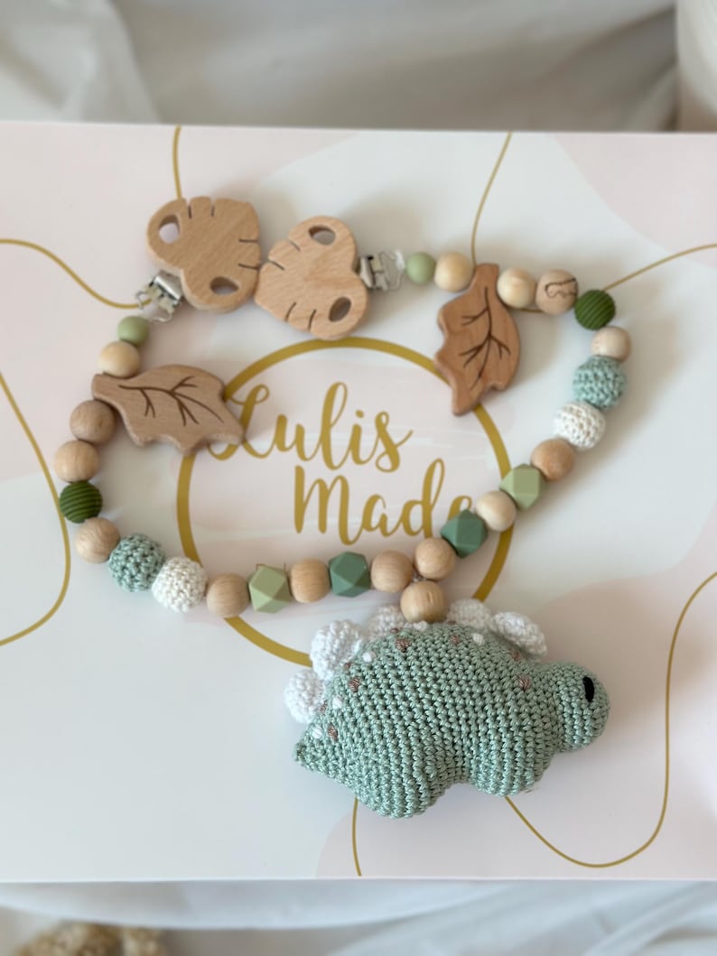 May include: A baby pacifier clip with a green crocheted dinosaur, wooden beads, and leaf accents. The clip features a wooden butterfly-shaped clip and the text "Lulis Made". The color palette includes shades of green, white, and natural wood.
