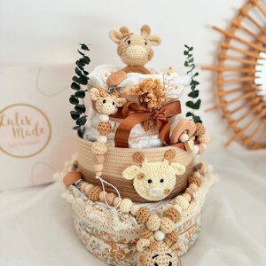 K&ouml;nnte beinhalten: Ein dreistufiger Babygeschenkkorb mit Giraffenmotiv. Der Korb hat geh&auml;kelte Giraffenakzente, Holzperlen und ein braunes Band. Die untere Ebene hat einen Stoff mit Blumenmuster. Die obere Ebene hat eine geh&auml;kelte Giraffe und einen Schnullerclip.