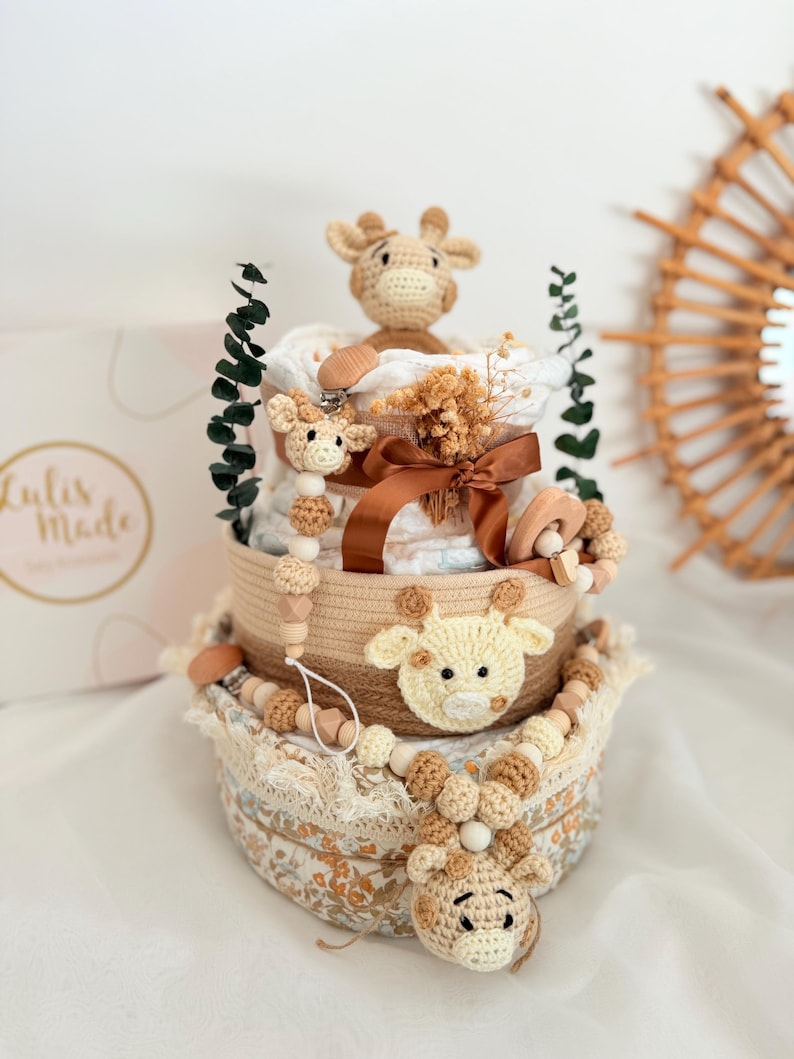 K&ouml;nnte beinhalten: Ein dreistufiger Babygeschenkkorb mit Giraffenmotiv. Der Korb hat geh&auml;kelte Giraffenakzente, Holzperlen und ein braunes Band. Die untere Ebene hat einen Stoff mit Blumenmuster. Die obere Ebene hat eine geh&auml;kelte Giraffe und einen Schnullerclip.