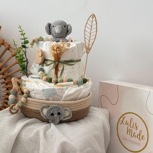 Op de afbeelding: Een luiercake versierd met een grijze gehaakte olifant, houten kralen en een groene bladertak. De cake staat in een geweven mand met een grijze gehaakte olifant aan de onderkant. De cake staat op een wit oppervlak met een doos waarop "Lulis Made Baby Accessories" staat.