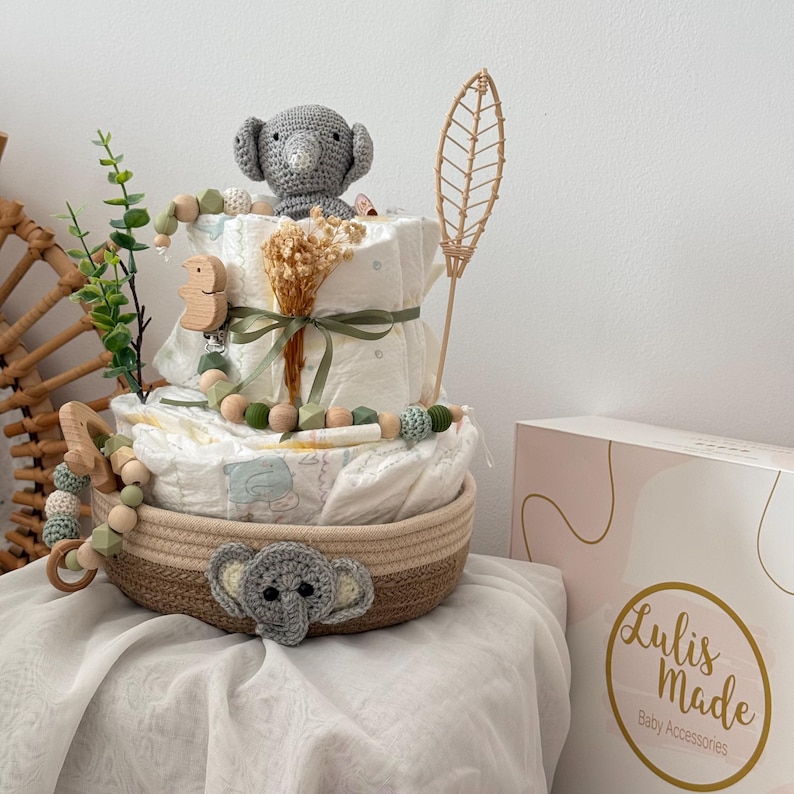 Op de afbeelding: Een luiercake versierd met een grijze gehaakte olifant, houten kralen en een groene bladertak. De cake staat in een geweven mand met een grijze gehaakte olifant aan de onderkant. De cake staat op een wit oppervlak met een doos waarop "Lulis Made Baby Accessories" staat.