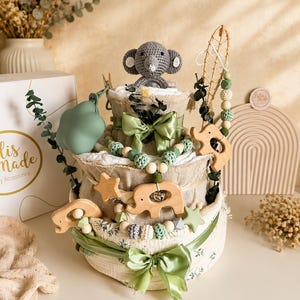 May include: A three-tiered baby gift basket with a gray crocheted elephant topper. The basket is decorated with green ribbons, wooden elephant and star ornaments, and a beaded garland. A green pacifier and a box with gold text are also visible.