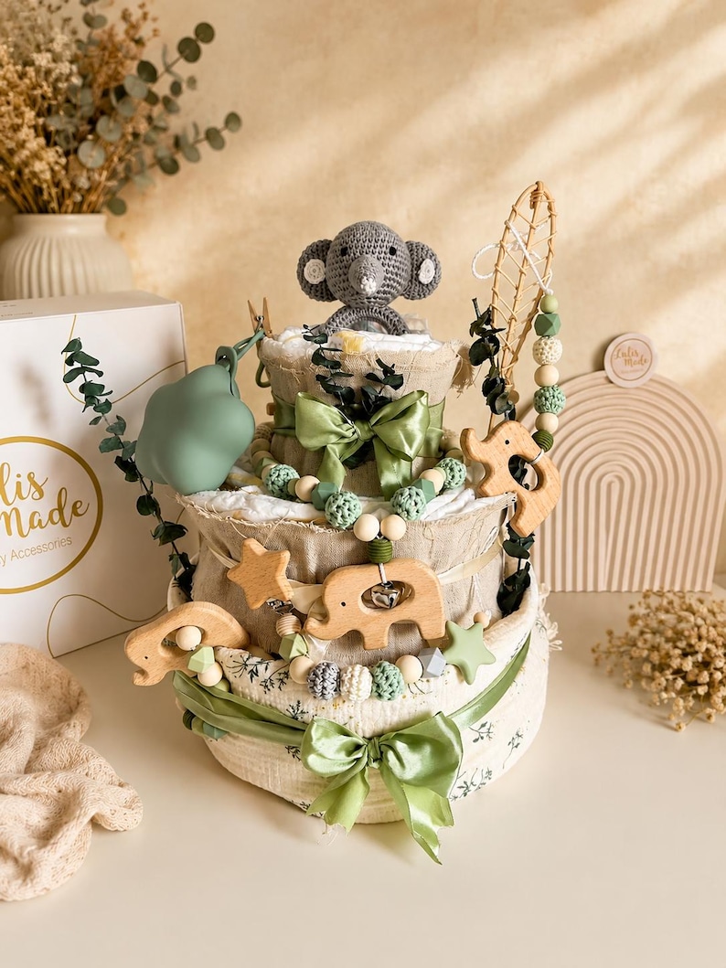 May include: A three-tiered baby gift basket with a gray crocheted elephant topper. The basket is decorated with green ribbons, wooden elephant and star ornaments, and a beaded garland. A green pacifier and a box with gold text are also visible.