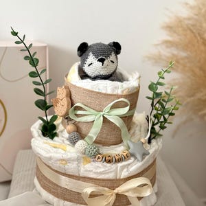 Peut inclure: Un g&acirc;teau &agrave; couches &agrave; deux &eacute;tages d&eacute;cor&eacute; d'un animal en crochet gris, de perles en bois et d'une &eacute;toile. Le g&acirc;teau est envelopp&eacute; de toile de jute et nou&eacute; avec un ruban vert et cr&egrave;me. L'&eacute;tage inf&eacute;rieur porte une &eacute;tiquette de nom en bois qui dit "PAUL".