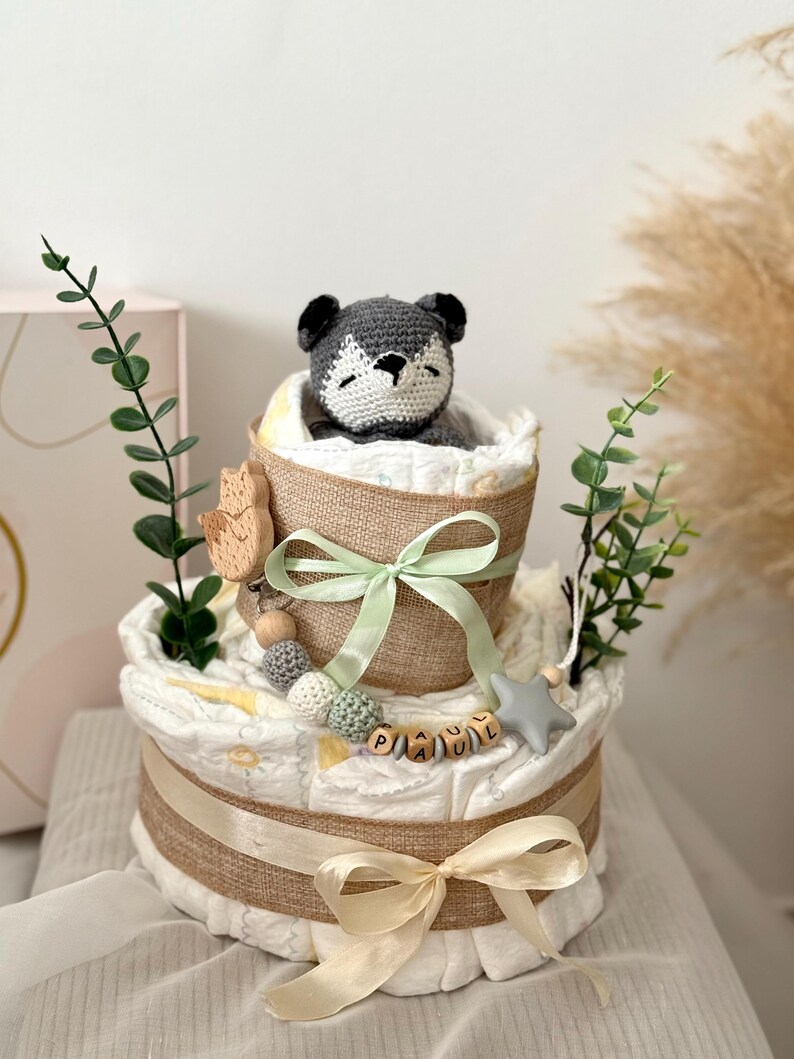 Peut inclure: Un g&acirc;teau &agrave; couches &agrave; deux &eacute;tages d&eacute;cor&eacute; d'un animal en crochet gris, de perles en bois et d'une &eacute;toile. Le g&acirc;teau est envelopp&eacute; de toile de jute et nou&eacute; avec un ruban vert et cr&egrave;me. L'&eacute;tage inf&eacute;rieur porte une &eacute;tiquette de nom en bois qui dit "PAUL".