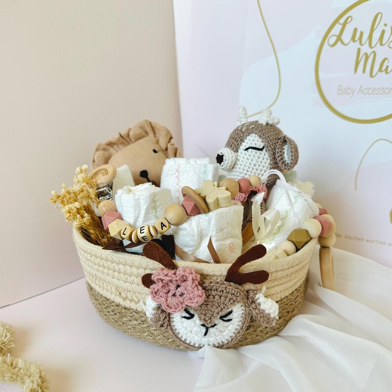 May include: A brown wicker basket filled with baby items, including a crocheted deer toy, a lion stuffed animal, and a wooden bead necklace with the name "LEA" spelled out. The basket is decorated with a crocheted deer head with pink flowers.