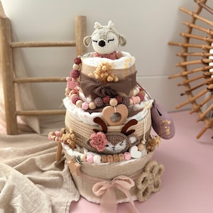 May include: A three-tiered baby shower gift basket with a crocheted deer toy on top. The basket is decorated with a pink ribbon, wooden beads, and a brown burlap bow. The basket is filled with baby items, such as diapers, wipes, and toys.