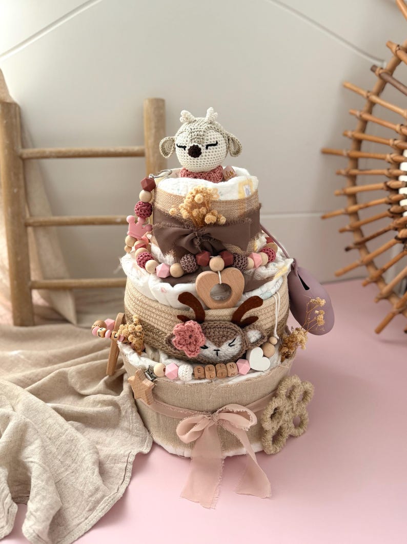 May include: A three-tiered baby shower gift basket with a crocheted deer toy on top. The basket is decorated with a pink ribbon, wooden beads, and a brown burlap bow. The basket is filled with baby items, such as diapers, wipes, and toys.
