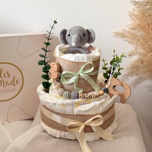 Peut inclure: Un g&acirc;teau &agrave; couches &agrave; deux &eacute;tages d&eacute;cor&eacute; d'un &eacute;l&eacute;phant en crochet gris, de feuilles vertes, d'un hochet en bois et d'un ruban beige. Le g&acirc;teau est envelopp&eacute; de toile de jute et porte une &eacute;tiquette de nom en bois personnalis&eacute;e qui dit "VIELY".