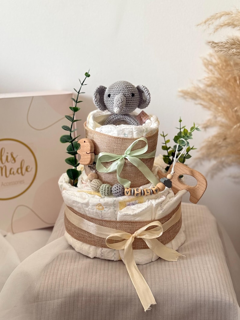Peut inclure: Un g&acirc;teau &agrave; couches &agrave; deux &eacute;tages d&eacute;cor&eacute; d'un &eacute;l&eacute;phant en crochet gris, de feuilles vertes, d'un hochet en bois et d'un ruban beige. Le g&acirc;teau est envelopp&eacute; de toile de jute et porte une &eacute;tiquette de nom en bois personnalis&eacute;e qui dit "VIELY".