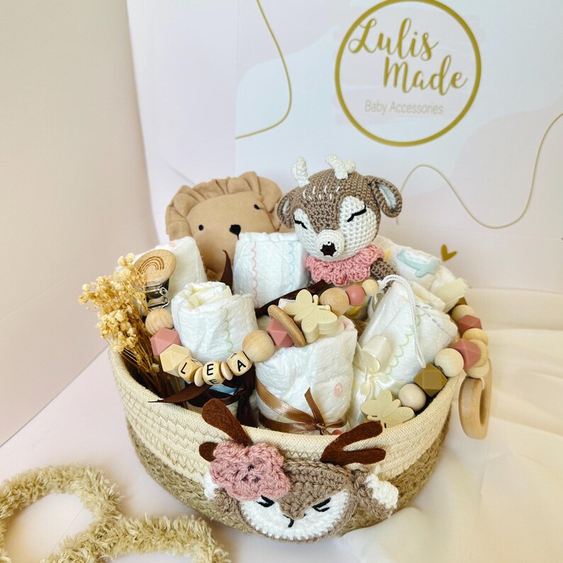 May include: A brown wicker basket filled with baby gifts, including a crocheted deer toy, a lion plush, wooden beads, and a white muslin cloth with the name "LEA" spelled out in wooden letters. The basket is decorated with a crocheted deer head.