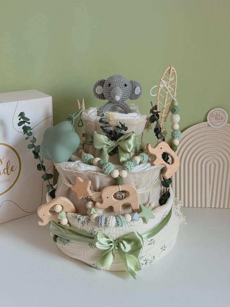 May include: A three-tiered baby diaper cake with a gray crocheted elephant topper. The cake is decorated with green ribbons, wooden elephant and star accents, and a beaded garland. A box with the text "Lulis Made" is visible in the background.