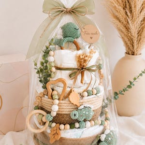May include: A diaper cake gift wrapped in clear cellophane and tied with a green ribbon. The cake is decorated with a green crochet dinosaur, wooden beads, and a wooden heart-shaped toy. A small wooden tag says "F&uuml;r euch".