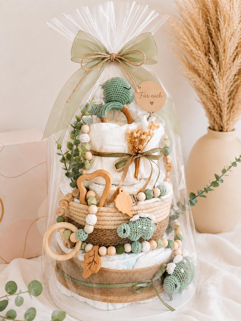 May include: A diaper cake gift wrapped in clear cellophane and tied with a green ribbon. The cake is decorated with a green crochet dinosaur, wooden beads, and a wooden heart-shaped toy. A small wooden tag says "F&uuml;r euch".
