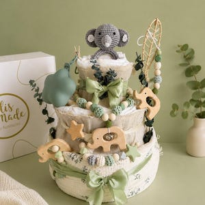 May include: A three-tiered baby diaper cake with a gray crocheted elephant topper. The cake is decorated with green ribbons, wooden elephant and star ornaments, and green and white beads. The cake sits next to a white box with gold text.