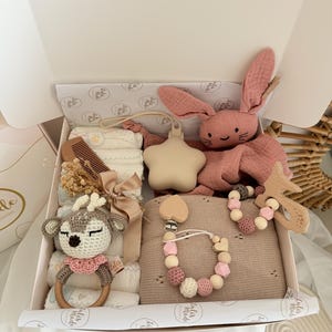 May include: A baby gift box filled with various items in soft, neutral tones. Includes a pink bunny plush, a wooden deer rattle, a star-shaped teether, a wooden comb, a blanket, and a pacifier clip with wooden beads.
