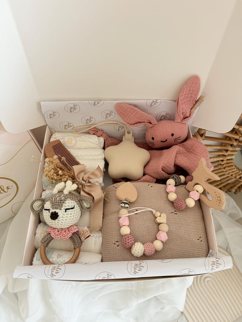 May include: A baby gift box filled with various items in soft, neutral tones. Includes a pink bunny plush, a wooden deer rattle, a star-shaped teether, a wooden comb, a blanket, and a pacifier clip with wooden beads.