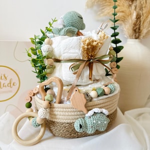 May include: A diaper cake with a woven basket base, decorated with a crocheted turtle, wooden beads, and greenery. The cake has two tiers of diapers, tied with a ribbon. A wooden teether and pacifier clip are also included.