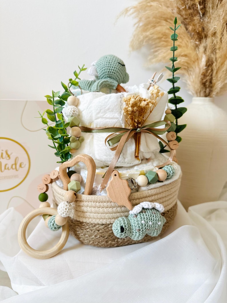 May include: A diaper cake with a woven basket base, decorated with a crocheted turtle, wooden beads, and greenery. The cake has two tiers of diapers, tied with a ribbon. A wooden teether and pacifier clip are also included.