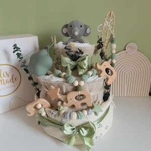 May include: A three-tiered baby diaper cake decorated with green bows, wooden elephant and star accents, and a crocheted gray elephant topper. The cake is adorned with green and white beads and a decorative leaf.