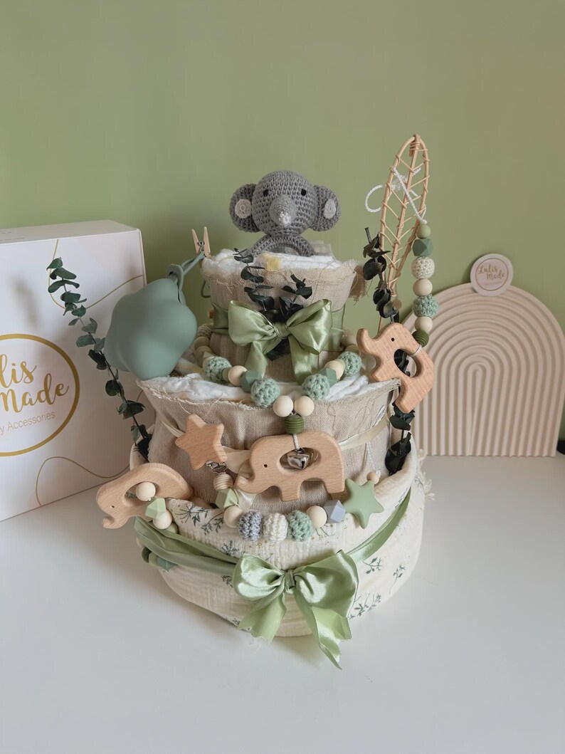 May include: A three-tiered baby diaper cake decorated with green bows, wooden elephant and star accents, and a crocheted gray elephant topper. The cake is adorned with green and white beads and a decorative leaf.