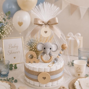 May include: A baby shower centerpiece featuring a diaper cake with a gray crochet elephant, wooden accents, and a bow. The cake is wrapped in cellophane and surrounded by balloons, flowers, and a sign that says "Baby Shower".