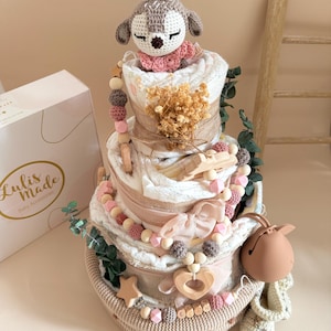May include: A multi-tiered baby diaper cake with a crochet deer topper, adorned with wooden beads, a star, and a heart. The cake is decorated with pink and beige accents, greenery, and a box that says "Lulis Made Baby Accessories."