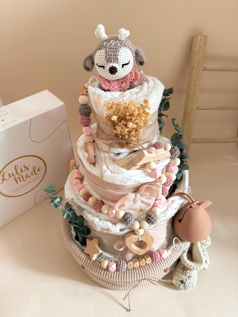 May include: A multi-tiered baby diaper cake with a crochet deer topper, adorned with wooden beads, a star, and a heart. The cake is decorated with pink and beige accents, greenery, and a box that says "Lulis Made Baby Accessories."