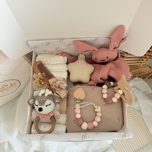May include: A baby gift box filled with a variety of items. Includes a pink bunny plush, a star-shaped rattle, a wooden comb, a crocheted deer rattle, a pacifier clip, and a blanket. The box is white with a decorative interior.