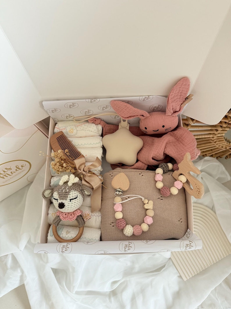 May include: A baby gift box filled with a variety of items. Includes a pink bunny plush, a star-shaped rattle, a wooden comb, a crocheted deer rattle, a pacifier clip, and a blanket. The box is white with a decorative interior.