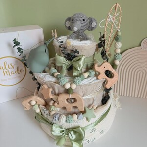 May include: A three-tiered baby gift set resembling a cake, featuring a gray crocheted elephant, wooden animal shapes, and sage green ribbons. The set includes a box with the text "ulis made Baby Accessories".