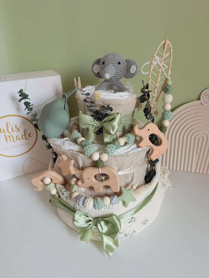 May include: A three-tiered baby gift set resembling a cake, featuring a gray crocheted elephant, wooden animal shapes, and sage green ribbons. The set includes a box with the text "ulis made Baby Accessories".