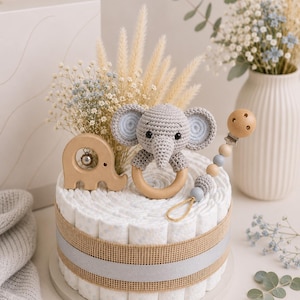 May include: A diaper cake decorated with a gray crocheted elephant toy, wooden elephant rattle, and a pacifier clip. The cake is wrapped with a burlap ribbon and a gray ribbon. The cake is decorated with dried flowers and baby's breath.