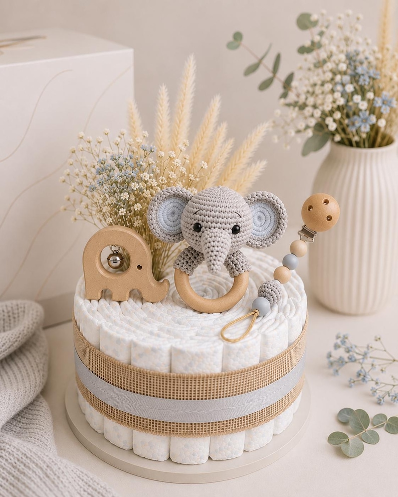 May include: A diaper cake decorated with a gray crocheted elephant toy, wooden elephant rattle, and a pacifier clip. The cake is wrapped with a burlap ribbon and a gray ribbon. The cake is decorated with dried flowers and baby's breath.
