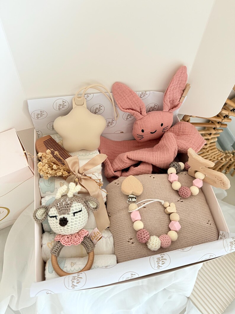 May include: A baby gift box filled with a variety of items. Includes a beige star-shaped teether, a pink plush bunny, a crochet deer rattle, a wooden comb, a pacifier clip, and a knitted blanket. The box is white with a pink interior.
