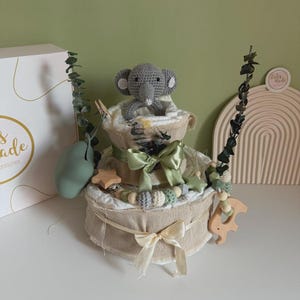 May include: A two-tiered baby gift basket with a gray crocheted elephant topper. The basket is decorated with green bows, wooden stars, and a wooden horse-shaped teether. The basket is wrapped in beige fabric.