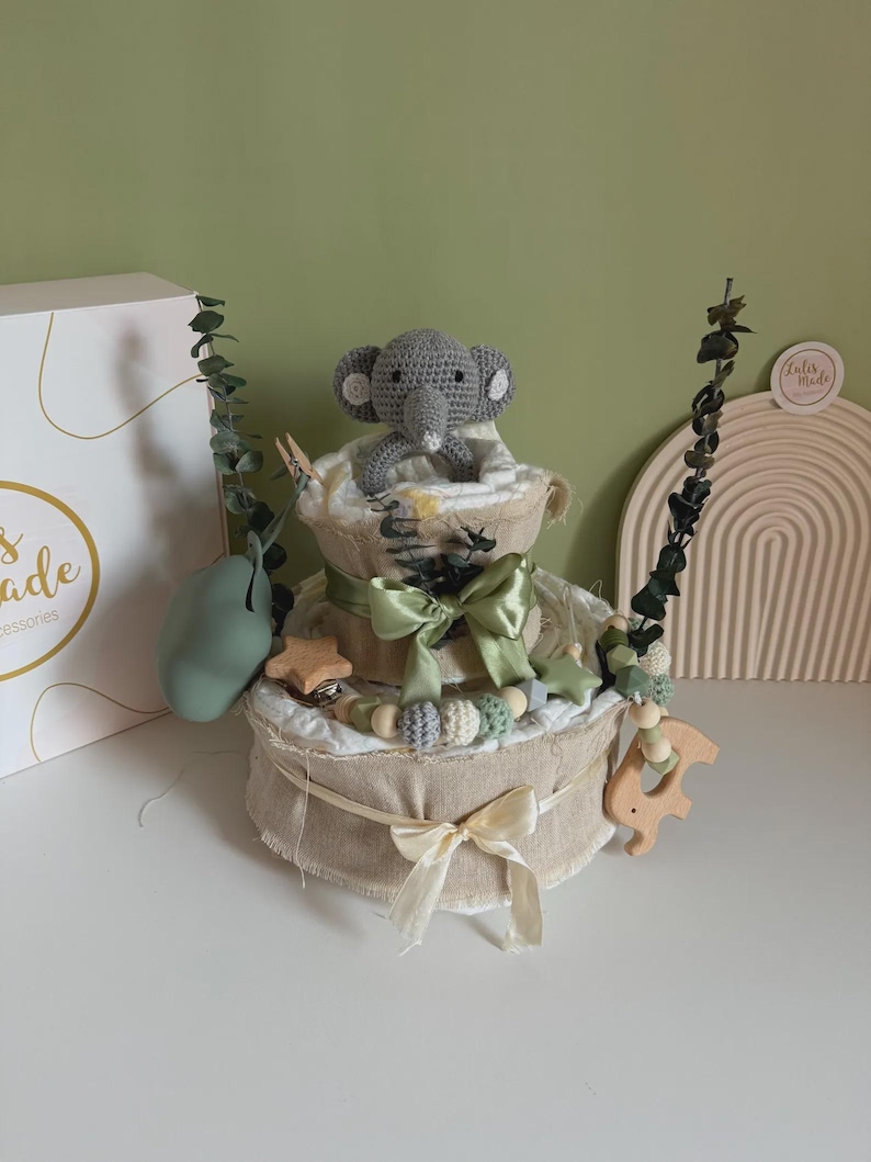 May include: A two-tiered baby gift basket with a gray crocheted elephant topper. The basket is decorated with green bows, wooden stars, and a wooden horse-shaped teether. The basket is wrapped in beige fabric.